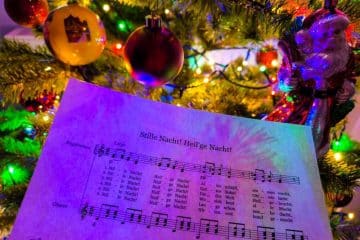 Ein Notenblatt für Stille Nacht! Heilige Nacht! (Stille Nacht) wird vor einem geschmückten Weihnachtsbaum mit bunten Lichtern, Ornamenten und einer Weihnachtsmannfigur gehalten und erinnert an den Charme klassischer japanischer Weihnachtslieder.