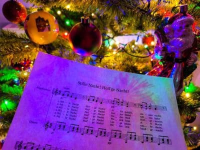 Ein Notenblatt für Stille Nacht! Heilige Nacht! (Stille Nacht) wird vor einem geschmückten Weihnachtsbaum mit bunten Lichtern, Ornamenten und einer Weihnachtsmannfigur gehalten und erinnert an den Charme klassischer japanischer Weihnachtslieder.