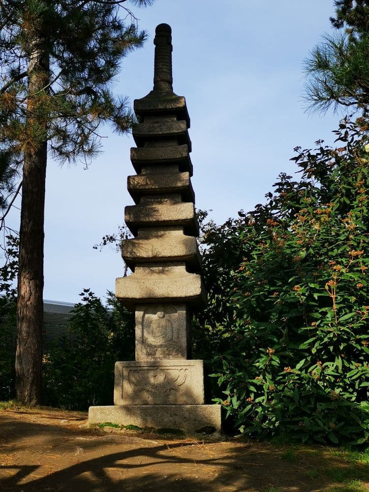 Eine hohe, mehrstöckige Steinpagode steht zwischen Bäumen und Sträuchern im Japanischen Garten im Nordpark Düsseldorf. Sie wird vom Sonnenlicht angestrahlt und erinnert an eine heitere japanische Atmosphäre unter einem klaren Himmel.