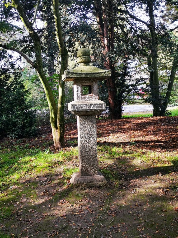 Eine traditionelle Steinlaterne steht auf einer Rasenfläche unter Bäumen in einem Japanischen Garten und wirft einen kleinen Schatten im Sonnenlicht. Bäume und Sträucher füllen den Hintergrund und evozieren eine ruhige japanische Atmosphäre im Nordpark Düsseldorf.