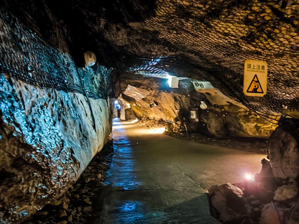 Ein schwach beleuchteter unterirdischer Tunnel mit felsigen Wänden, Netzen über dem Boden, Warnschildern und Lichtern, die den Weg beleuchten. Der Iwaya-Tunnel erscheint feucht und hat einen glatten Betonweg.
