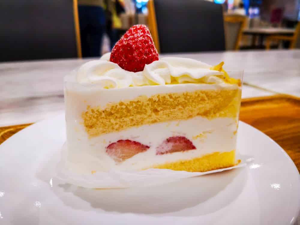 Ein Stück Erdbeerkuchen, inspiriert von der japanischen Kurisumasukēki-Tradition, mit Schichten aus Biskuit, Schlagsahne und Erdbeeren, gekrönt mit einer ganzen Erdbeere, serviert auf einem weißen Teller in einem gemütlichen Café.