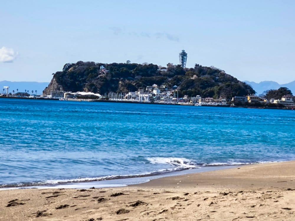 Ein Sandstrand führt zu ruhigem, blauem Wasser mit einer baumbewachsenen Insel und dem ikonischen Aussichtsturm Enoshima Sea Candle, der sich in der Ferne unter einem klaren blauen Himmel erhebt.