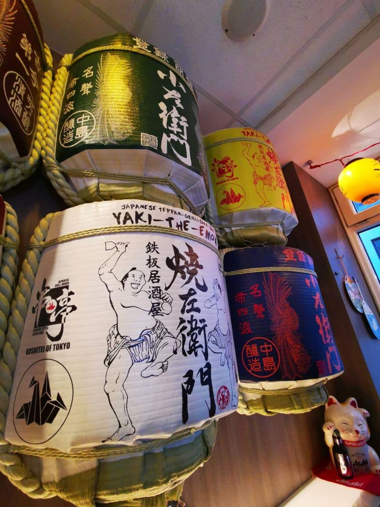 Große dekorative Sake-Fässer mit japanischen Schriftzügen und farbenfrohen Mustern sind an einer Wand in Little Tokyo, Düsseldorf, ausgestellt. Auf einem Fass ist ein Sumo-Ringer abgebildet, während darunter eine Maneki-neko-Figur (winkende Katze) sitzt.