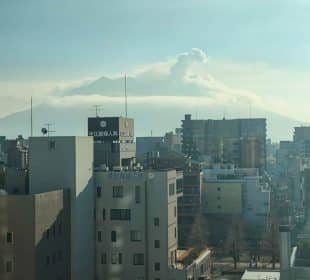 Die Gebäude der Stadt im Vordergrund und der große, teilweise verdeckte Vulkan von Kagoshima im Hintergrund, der eine Rauchfahne ausstößt - einer von vielen Gründen, diese Stadt auf Ihre Reiseliste zu setzen - alles unter einem dunstigen blauen Himmel.