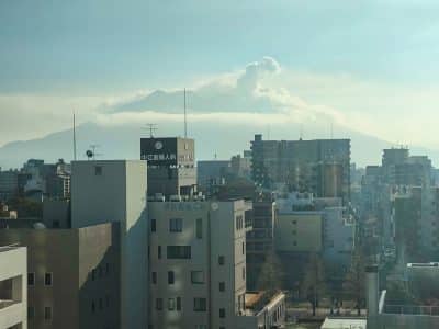Die Gebäude der Stadt im Vordergrund und der große, teilweise verdeckte Vulkan von Kagoshima im Hintergrund, der eine Rauchfahne ausstößt - einer von vielen Gründen, diese Stadt auf Ihre Reiseliste zu setzen - alles unter einem dunstigen blauen Himmel.