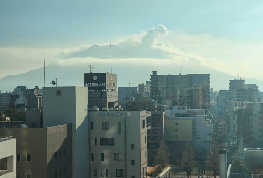 Die Gebäude der Stadt im Vordergrund und der große, teilweise verdeckte Vulkan von Kagoshima im Hintergrund, der eine Rauchfahne ausstößt - einer von vielen Gründen, diese Stadt auf Ihre Reiseliste zu setzen - alles unter einem dunstigen blauen Himmel.