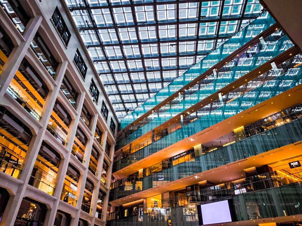 Ein modernes Atrium in Marunouchi mit mehreren Etagen, Glasgeländern und einer großen Glasdecke, die natürliches Licht hereinlässt, schafft Reflexionen und eine offene, helle Atmosphäre. Das Gebäude zeichnet sich durch warme Beleuchtung und modernes Design aus.