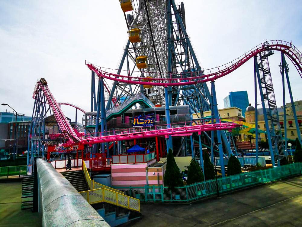 Eine lebhafte Cosmo World-Vergnügungsparkszene mit einer rosafarbenen Achterbahn, die sich um ein großes Riesenrad dreht, umgeben von Gebäuden und Grünflächen unter einem strahlenden Himmel.