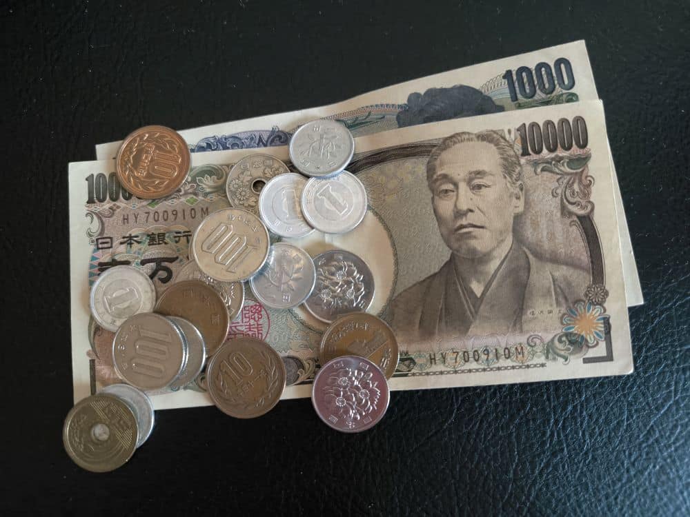 Japanische Yen-Scheine und verschiedene Münzen sind auf einer schwarzen Fläche ausgebreitet, als ob jemand sein Geld verloren hätte. Es handelt sich um Scheine im Wert von 10.000 und 1.000 Yen und um Münzen mit verschiedenen Werten, die verstreut sind.