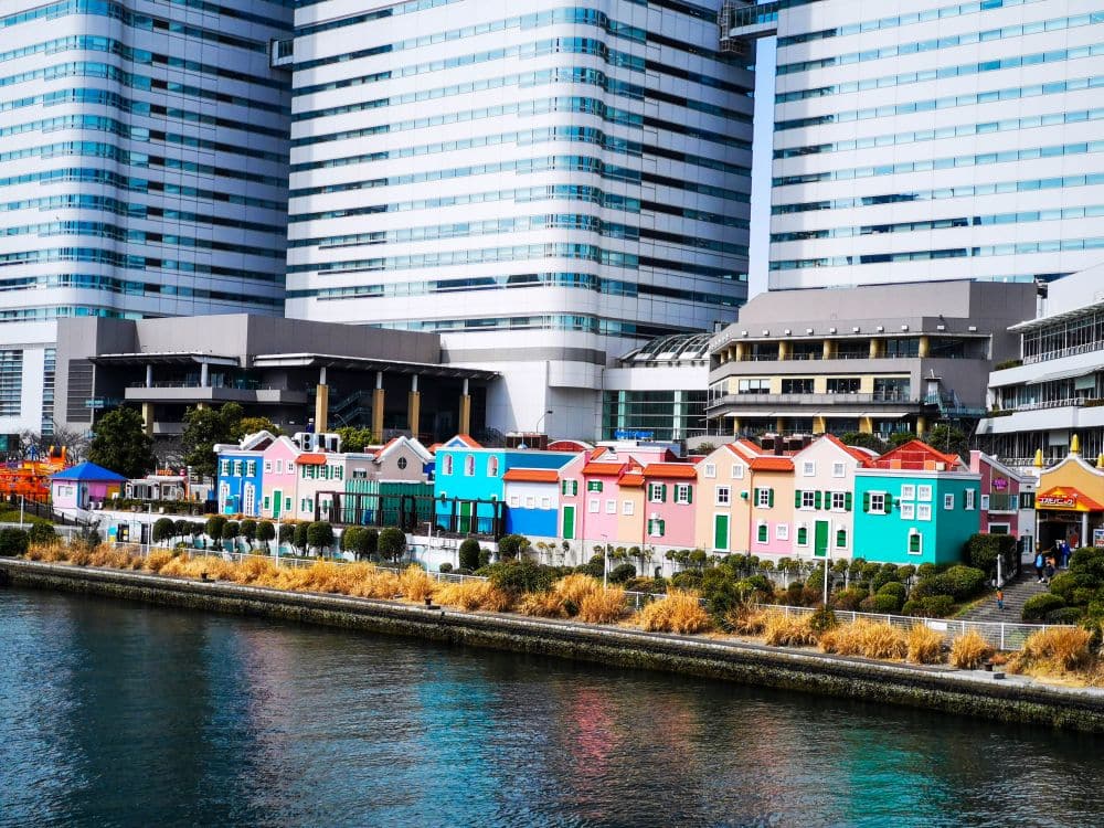 Eine Reihe bunter Häuser steht am Wasser in der Nähe von Cosmo World, mit modernen, hohen Bürogebäuden im Hintergrund und etwas Grün entlang des Ufers.
