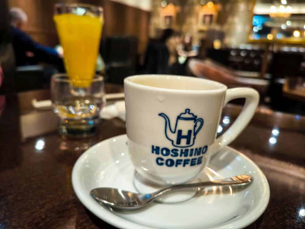 Eine weiße Kaffeetasse mit dem Aufdruck "Hoshino Coffee" steht auf einer Untertasse mit einem Löffel auf einem dunklen Tisch in einem gemütlichen Café in Marunouchi. Im Hintergrund stehen ein Glas Orangensaft und Leute, die sitzen und sich unterhalten.
