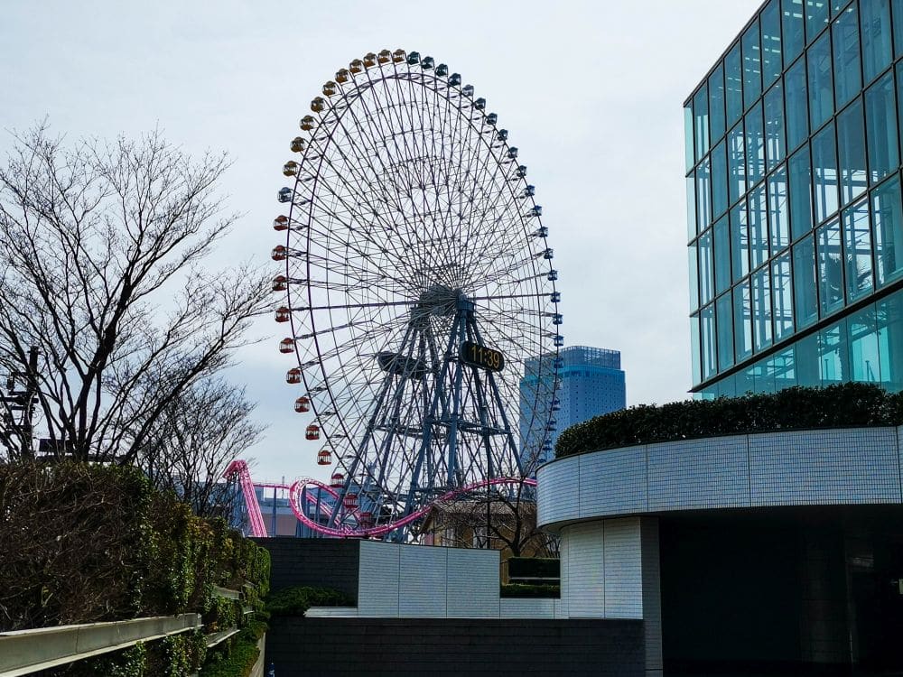Ein großes Riesenrad mit bunten Gondeln, Teil von Cosmo World, steht an einem bewölkten Tag in der Nähe von Glasgebäuden und blattlosen Bäumen. Vor dem Rad schlängelt sich eine rosafarbene Achterbahnstrecke.