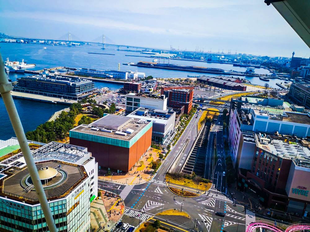 Luftaufnahme einer Küstenstadt mit modernen Gebäuden, Grünflächen, dem nahe gelegenen Vergnügungspark Cosmo World, einem großen Hafen, mehreren angedockten Schiffen und einer langen Brücke, die sich unter blauem Himmel über das Wasser spannt.