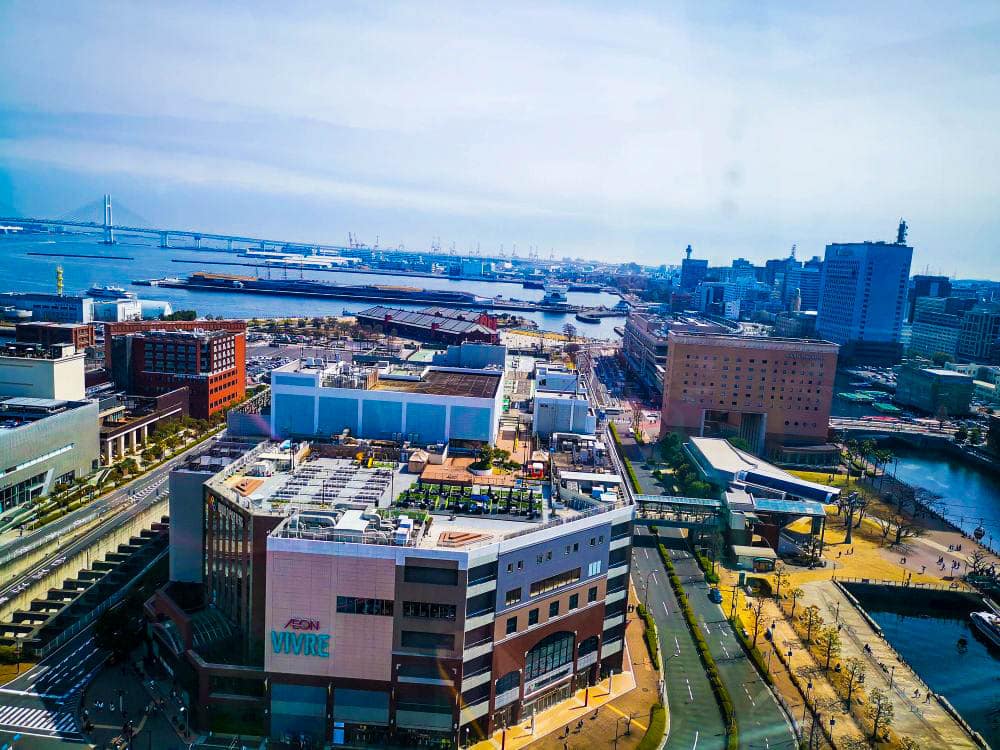 Luftaufnahme einer Küstenstadt mit modernen Gebäuden, Straßen und Cosmo World in der Ferne. Der Ozean erstreckt sich hinter der Stadtlandschaft, während eine Brücke den Horizont unter einem klaren blauen Himmel überspannt.