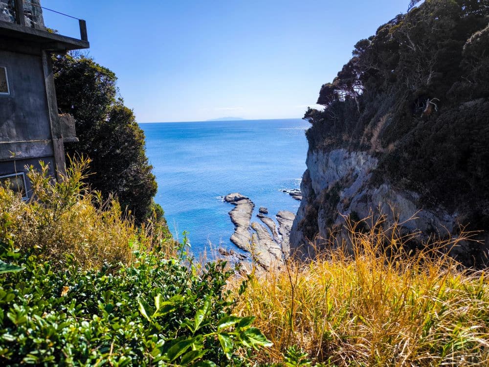 Ein Blick auf das blaue Meer, eingerahmt von felsigen Klippen und grünen Büschen, mit dem Daishi-Gebäude auf der linken Seite und hohem Gras im Vordergrund unter einem klaren, sonnigen Himmel.