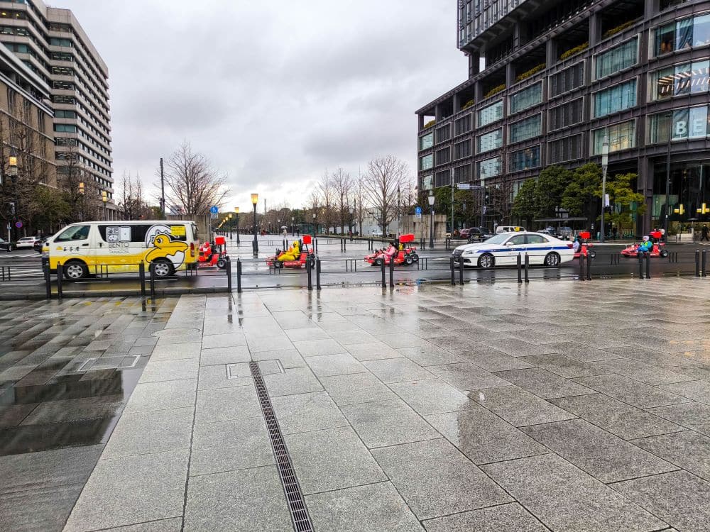 Eine Gruppe rot gekleideter Menschen fährt mit Go-Karts durch Marunouchi, gefolgt von einem Polizeiauto und einem gelben Lieferwagen mit einem Cartoon-Gesicht auf einer nassen, von modernen Gebäuden gesäumten Straße an einem bewölkten Tag.