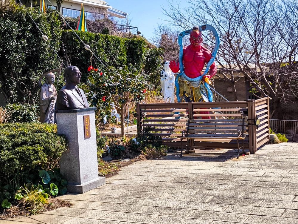 Eine Gartenszene mit einer Büstenstatue auf einem Sockel im Vordergrund und einer großen roten dämonenähnlichen Daishi-Statue mit blauen Flügeln und einem Schwert im Hintergrund, umgeben von Grün und einem Holzzaun.