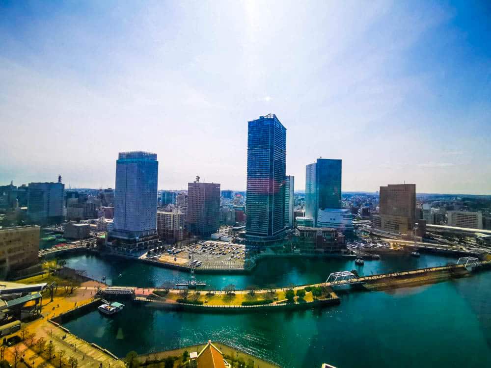 Eine pulsierende Stadtlandschaft mit hohen modernen Wolkenkratzern und Gebäuden, die einen Fluss oder Kanal umgeben, unter einem hellen, blauen Himmel. Das Sonnenlicht spiegelt sich auf dem Wasser und dem Riesenrad Cosmo World und hebt die dynamische städtische Umgebung hervor.