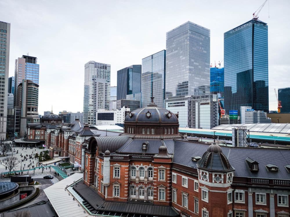 Ein Blick auf das historische rote Backsteingebäude des Tokioter Bahnhofs im Vordergrund, inmitten der modernen gläsernen Wolkenkratzer von Marunouchi unter einem bewölkten Himmel. Menschen spazieren in der Nähe des Bahnhofs und auf den Straßen der Stadt darunter.