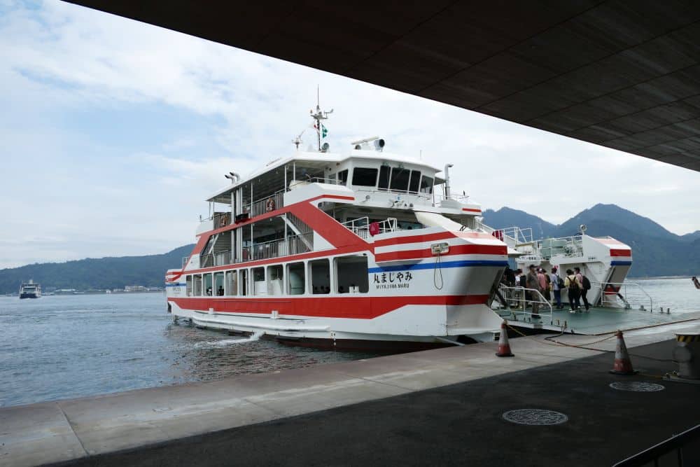 Eine weiß-rote Fähre liegt an einer Anlegestelle, wo Menschen an Bord gehen. Eine andere Fähre und Berge erscheinen im Hintergrund unter einem teilweise bewölkten Himmel.