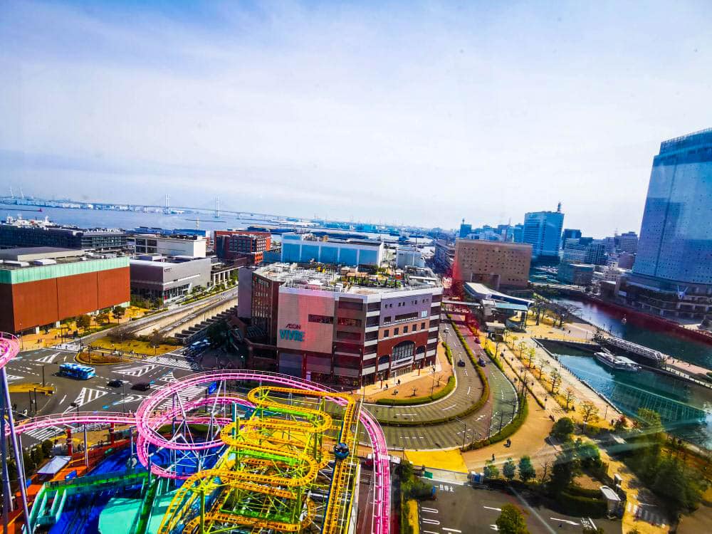 Eine lebendige Stadtlandschaft mit den bunten Achterbahnschienen von Cosmo World im Vordergrund, umgeben von modernen Gebäuden, breiten Straßen, einem Kanal und einem weiten Blick auf die Bucht unter einem klaren Himmel.