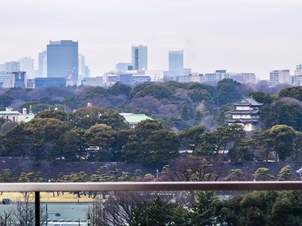 Blick auf das Stadtbild von Marunouchi mit modernen Wolkenkratzern im Hintergrund, einem dichten grünen Baumbestand in der Mitte und einem traditionellen japanischen Gebäude zwischen den Bäumen im Vordergrund.