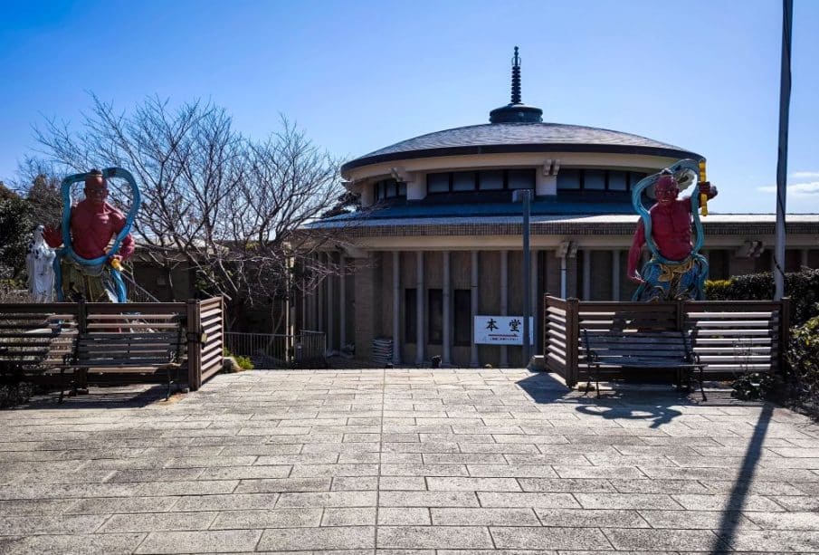 Im Hintergrund steht ein Gebäude mit rundem Dach und Säulen, flankiert von zwei großen, bunten Daishi-Wächterstatuen. Laublose Bäume und ein klarer blauer Himmel vervollständigen die Szene.