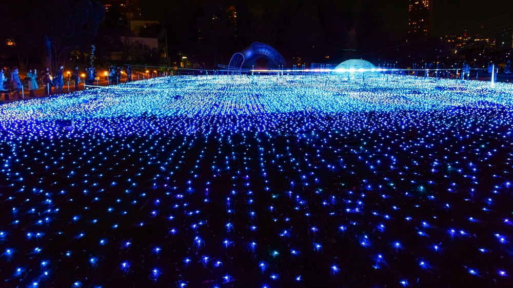 Ein großes Feld im Freien ist nachts mit Tausenden von leuchtenden blauen Lichtern bedeckt, die einen wellenartigen Effekt erzeugen. Die Gebäude und Bäume im Hintergrund werden von den Lichtern der Stadt sanft beleuchtet, was die magische Beleuchtung der Szene noch verstärkt.