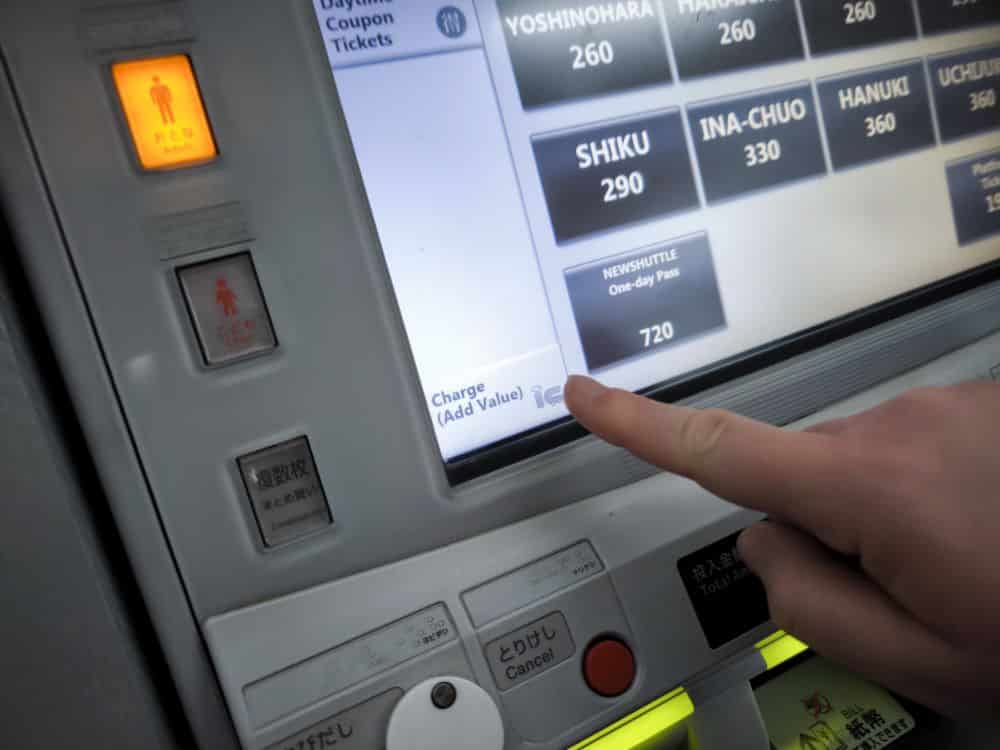 Eine Person zeigt mit dem Finger auf die Option Aufpreis (Add Value) auf einem Touchscreen-Fahrkartenautomaten, über dem verschiedene IC-Card-Fahrkartenoptionen und Preise angezeigt werden.