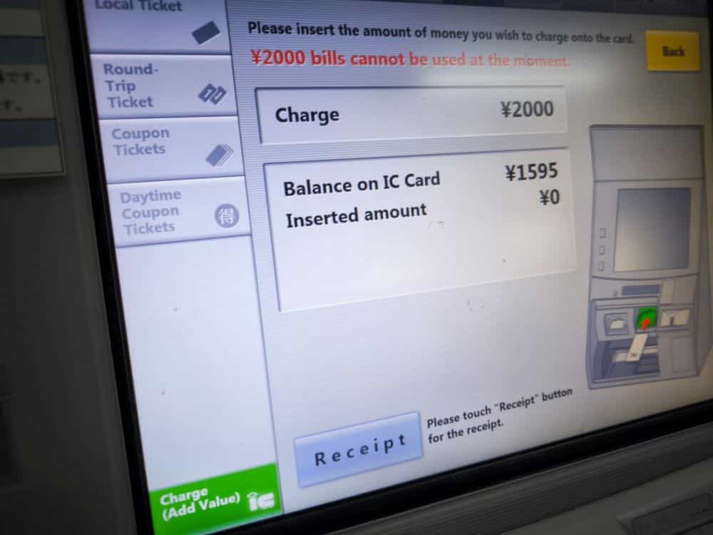 Der Bildschirm eines Fahrkartenautomaten zeigt eine Gebühr von ¥2000, ein aktuelles IC-Card-Guthaben von ¥1595 und einen Hinweis, dass ¥2000-Scheine nicht verwendet werden können. Optionen für die Art des Tickets, das Aufladen der IC-Karte und eine Quittungstaste sind sichtbar.