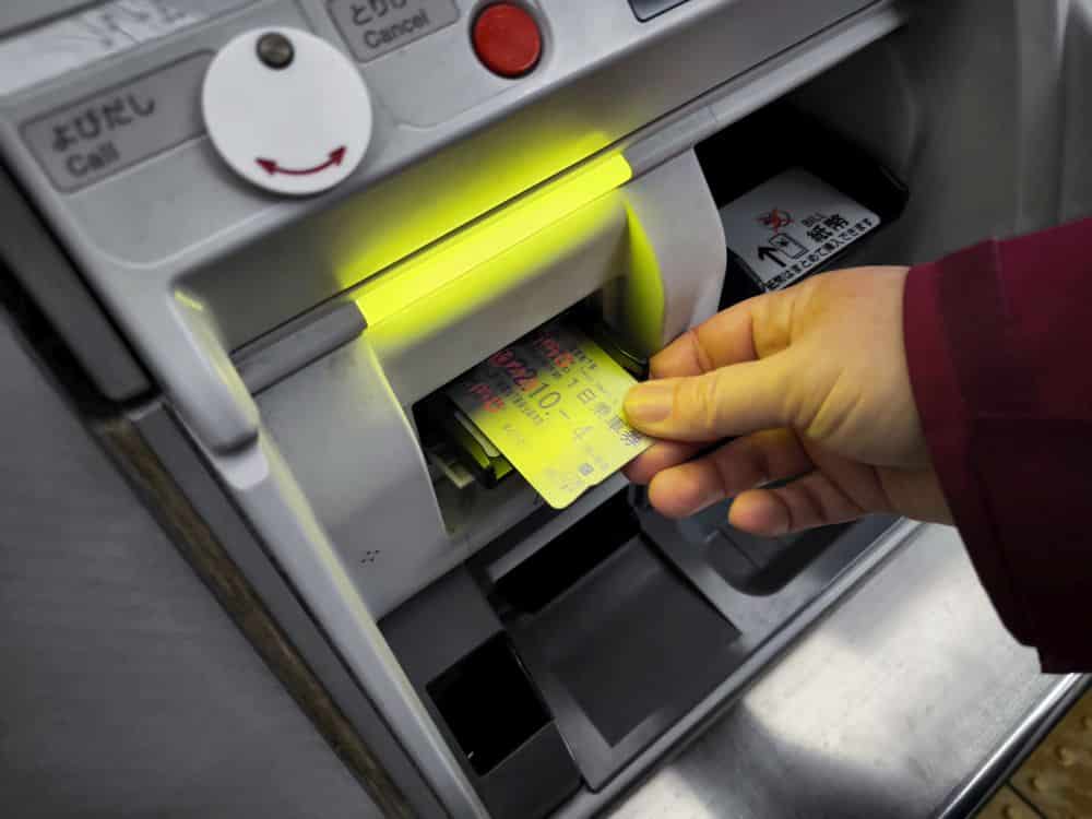 Eine Person steckt einen Fahrschein oder eine IC-Karte in einen Fahrkartenautomaten mit einem leuchtenden gelben Schlitz. Der Automat ist mit Tasten und japanischem Text auf dem Bedienfeld versehen.