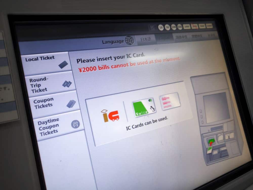 Bildschirm eines Geldautomaten mit Anweisungen zum Einführen einer IC-Karte; ein Hinweis besagt, dass ¥2000-Scheine nicht verwendet werden können. Auf der linken Seite sind die Ticketoptionen aufgelistet, und auf dem Bildschirm werden Symbole für verschiedene IC-Karten, einschließlich Suica, angezeigt.