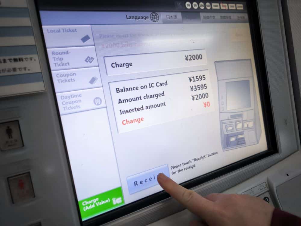 Nahaufnahme des Fingers einer Person, die die Quittungstaste auf dem Bildschirm eines Fahrkartenautomaten drückt, der die Zahlungsdaten in japanischen Yen anzeigt, mit Optionen für die Sprachauswahl, die Verwendung der IC-Karte und Informationen über den Kontostand der IC-Karte.