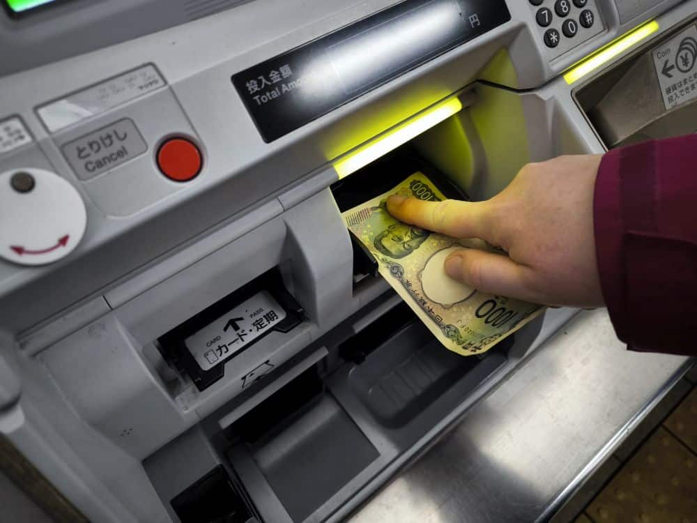 Eine Person in einem roten Ärmel steckt eine japanische 1000-Yen-Banknote in einen Geldautomaten, an dem Tasten, japanischer Text und ein IC-Kartenschlitz sichtbar sind.