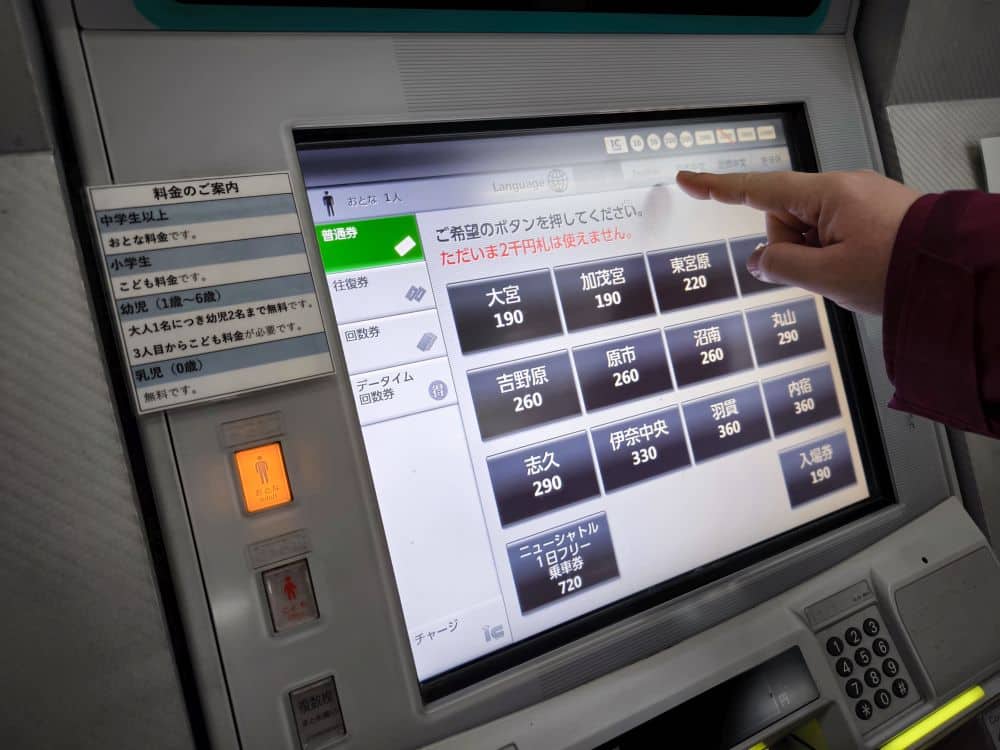 Die Hand einer Person berührt den Bildschirm eines japanischen Zugfahrkartenautomaten, auf dem verschiedene Fahrpreisbeträge, Fahrtzielnamen und IC-Kartenoptionen auf Japanisch angezeigt werden. Neben dem Bildschirm sind Schilder und Anweisungen zu sehen.