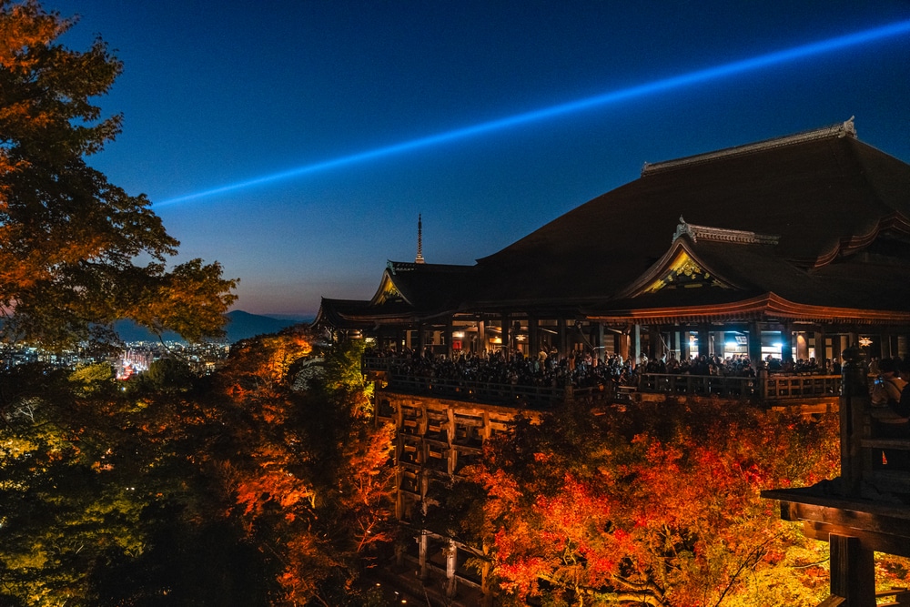 Der Kiyomizu-dera-Tempel in Kyoto, Japan, beleuchtet in der Abenddämmerung mit leuchtendem Herbstlaub, einer Menschenmenge auf der Terrasse und einem blauen Lichtstrahl, der sich über den Nachthimmel erstreckt.