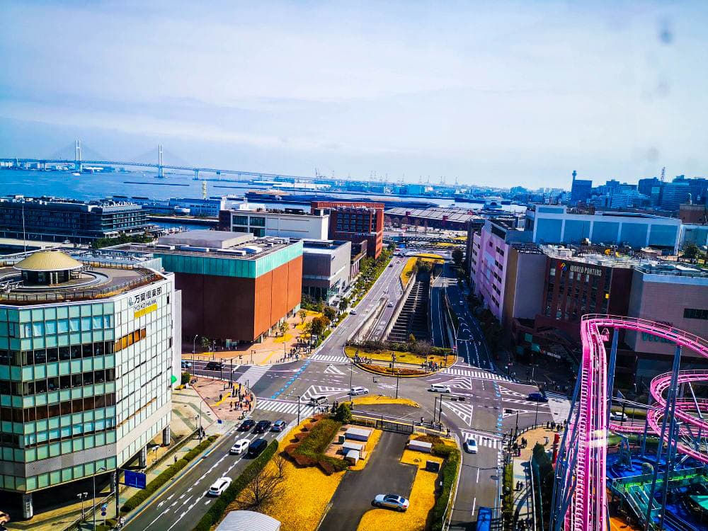 Ein pulsierendes Stadtbild mit modernen Gebäuden, sich kreuzenden Straßen, üppigem Grün und der legendären Cosmo World-Achterbahnstrecke. Im Hintergrund spannt sich eine große Brücke über das Wasser unter einem klaren Himmel.