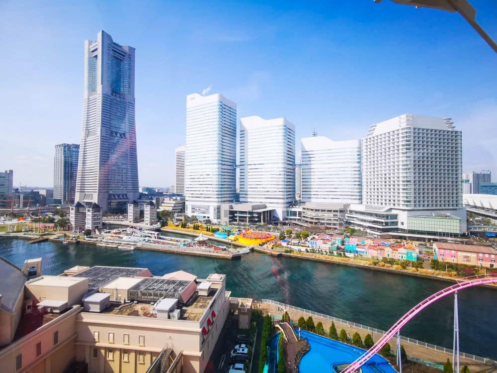 Eine Stadtansicht von Yokohama, Japan, mit modernen Hochhäusern, darunter der Landmark Tower, einem Kanal und dem Vergnügungspark Cosmo World mit seinen bunten Achterbahnschienen unter einem strahlend blauen Himmel.