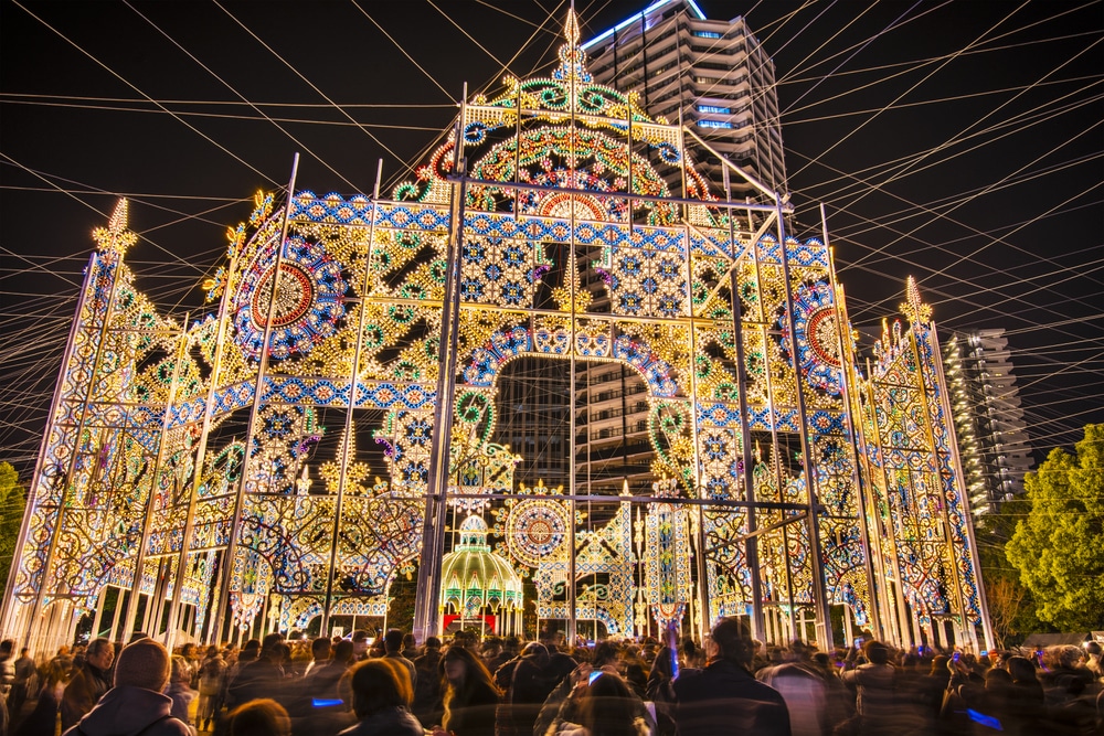 Eine große, verschnörkelte Beleuchtung mit verschlungenen Mustern und Bögen leuchtet nachts hell auf, während sich davor Menschenmengen versammeln und im Hintergrund hohe Gebäude aufragen.