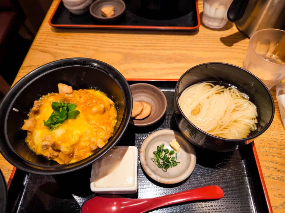 Eine japanische Mahlzeit in Marunouchi mit einer Schüssel Oyakodon (Huhn und Ei auf Reis), kalten Somen-Nudeln, Tofu, geschnittenen grünen Zwiebeln und Essiggurken, alles wunderschön auf einem schwarzen Tablett mit einem roten Löffel serviert.