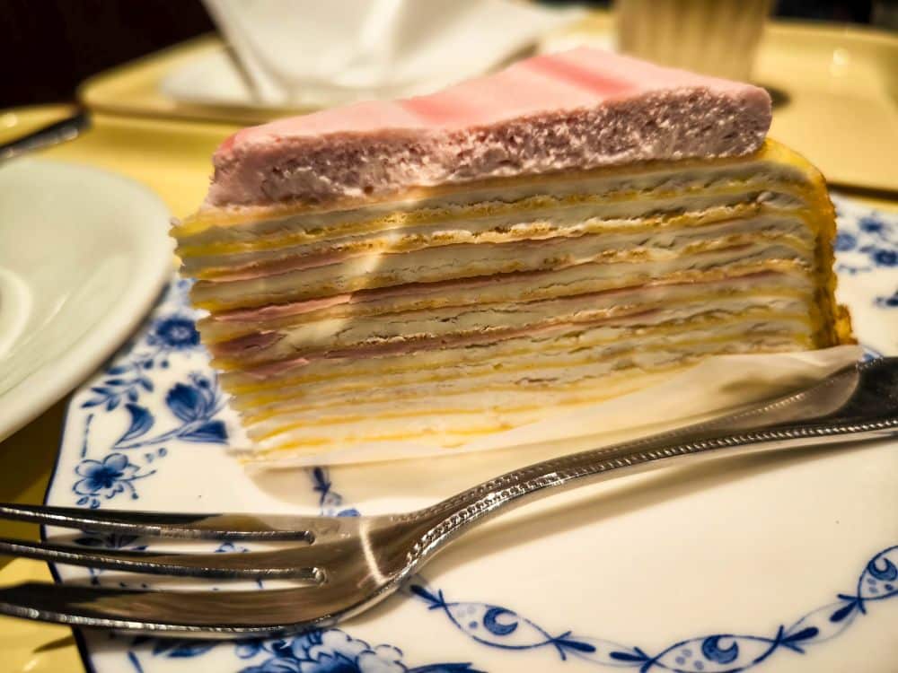 Nahaufnahme einer geschichteten Krepptorte mit rosa und weißen Cremeschichten, serviert auf einem dekorativen blau-weißen Teller in Marunouchi, mit einer Gabel daneben. Die Kuchenscheibe hat eine weiche, cremige Textur und eine pastellfarbene Oberfläche.