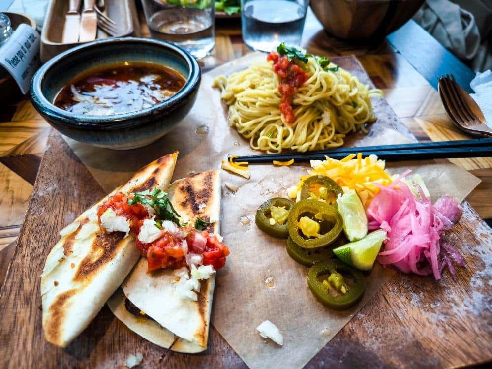 Ein Holzbrett mit Quesadillas mit Salsa, geschnittenen Jalapeños, eingelegten Zwiebeln, geriebenem Käse, Limettenspalten und einer Portion Marunouchi-Nudeln mit Tomatensoße, Stäbchen und einer Schale mit Brühe.