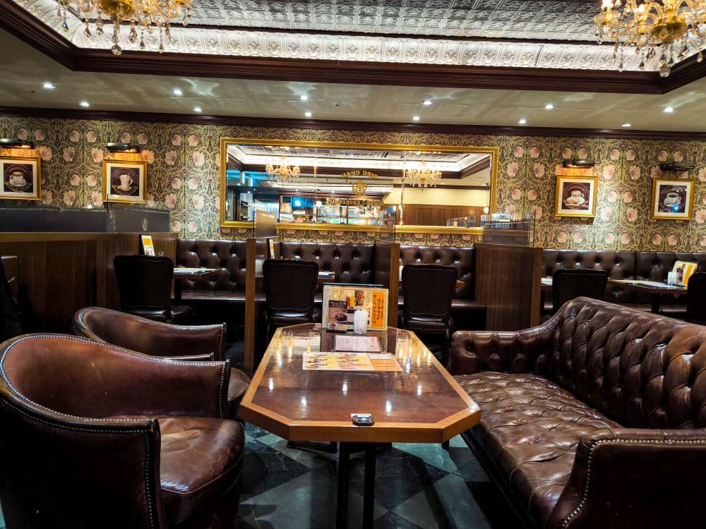 Ein gemütliches Restaurant im Vintage-Stil in Marunouchi mit braunen Ledersofas, Holztischen, gemusterten Tapeten, gerahmten Porträts an den Wänden und verschnörkelten Kronleuchtern, die von einer dekorativen Decke hängen.