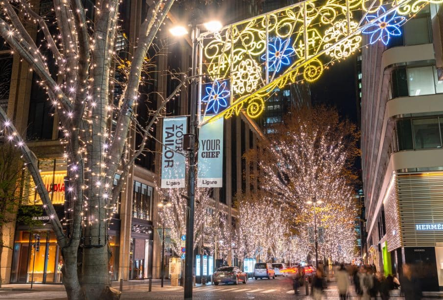 Eine nächtliche Stadtstraße, geschmückt mit funkelnden Lichtern an den Bäumen und einem bunten, beleuchteten Bogen über dem Kopf, während die Menschen an lebhaften Geschäften vorbeischlendern, die die hell erleuchtete Szene säumen. Illumination