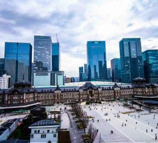 Luftaufnahme des historischen roten Backsteingebäudes des Tokioter Bahnhofs in Marunouchi, umgeben von modernen gläsernen Wolkenkratzern unter einem bewölkten Himmel, mit Menschen, die auf dem offenen Platz davor spazieren gehen.