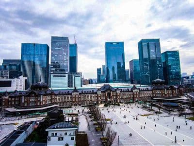 Luftaufnahme des historischen roten Backsteingebäudes des Tokioter Bahnhofs in Marunouchi, umgeben von modernen gläsernen Wolkenkratzern unter einem bewölkten Himmel, mit Menschen, die auf dem offenen Platz davor spazieren gehen.
