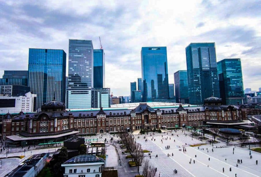 Luftaufnahme des historischen roten Backsteingebäudes des Tokioter Bahnhofs in Marunouchi, umgeben von modernen gläsernen Wolkenkratzern unter einem bewölkten Himmel, mit Menschen, die auf dem offenen Platz davor spazieren gehen.