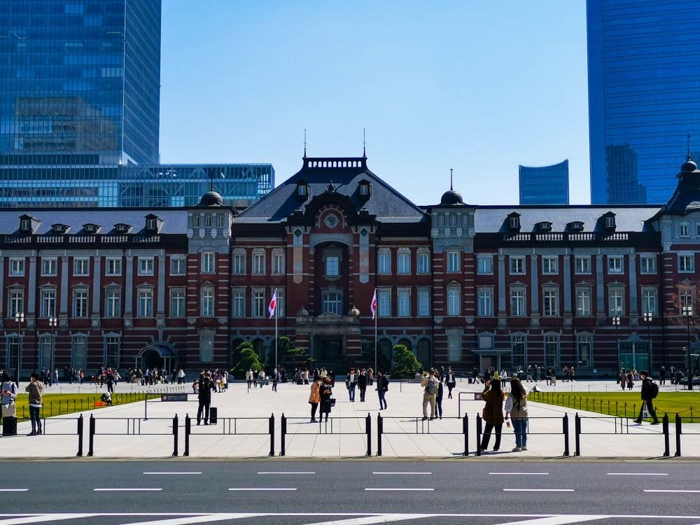 Die rote Backsteinfassade und die zentrale Kuppel des Tokioter Bahnhofs erheben sich stolz in Marunouchi, während in der Nähe Menschen spazieren gehen und sich im Hintergrund moderne Wolkenkratzer erheben. An diesem sonnigen Tag wehen zwei japanische Flaggen neben dem Eingang des Gebäudes.