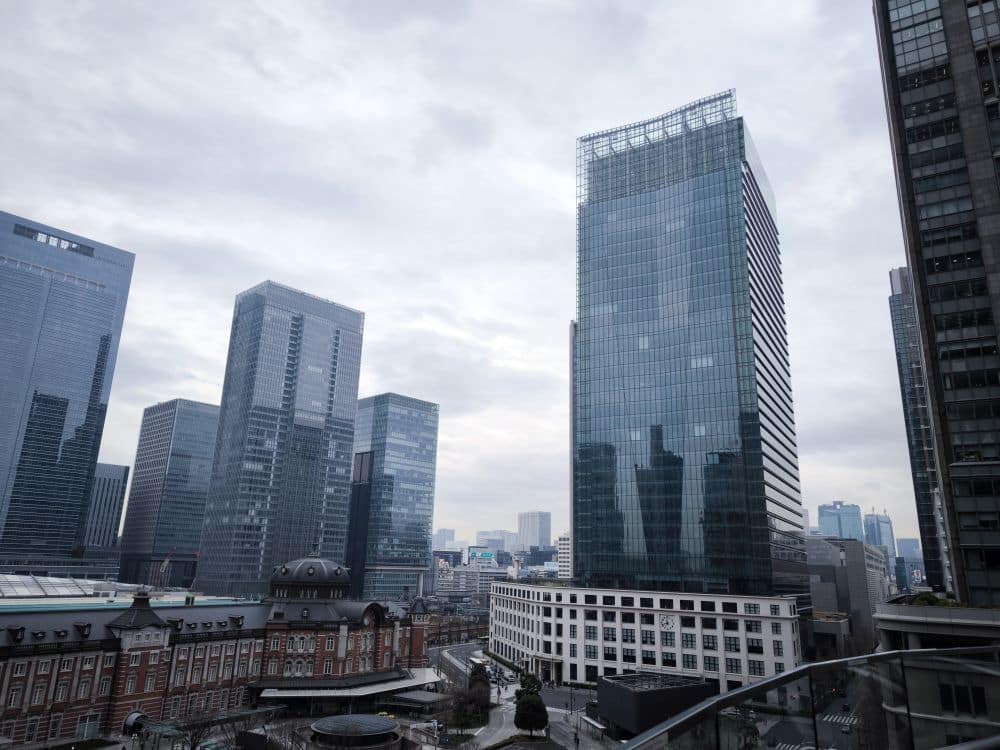 Eine Stadtansicht von Marunouchi mit modernen gläsernen Wolkenkratzern unter einem bewölkten Himmel, mit einem älteren, kuppelförmigen Gebäude im Vordergrund und mehreren Hochhäusern im Hintergrund.